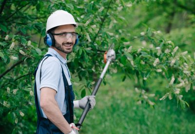 handsome gardener in helmet, protective glasses and noise-canceling headphones holding telescopic